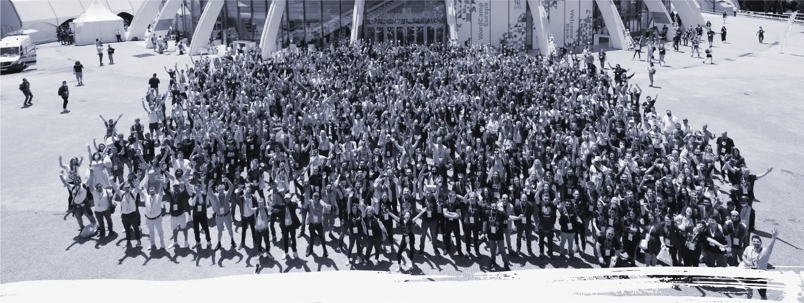 A black and white bird’s eye view of the thousands of contributors who attended WordCamp Europe 2022. They are all standing in front of the venue with their hands up in celebration.